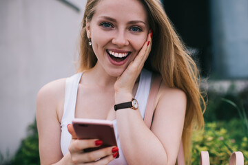 Half length portrait of stunned sincerely hipster girl smiling at camera and feeling expression emotions about winning mood, happy woman on city street using modern digital smartphone for phoning