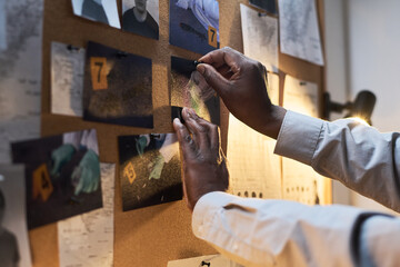 Hands pinning photos and notes on bulletin board in dimly lit room, with various pieces of evidence...