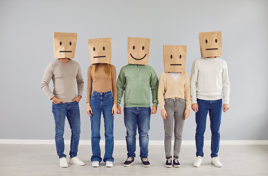 Happy smiley sanguine social man and group of apathetic phlegmatic indifferent people standing by grey wall background wearing brown shopping bags on heads with different drawn emoji mood expressions