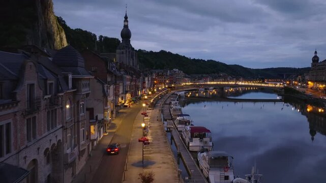Car glides along the Meuse river in Dinant, Belgium, as city lights shimmer on the water. Serene charm of dusk in this beautiful european town