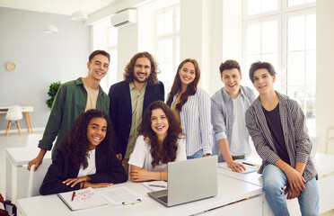 Group of high school students and classmates sitting at the desk with male friendly smiling teacher in modern classroom with laptop and looking at the camera. Education and back to school concept.