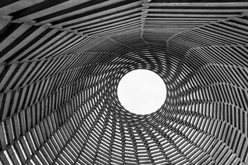 Up view of wooden beams spiralling upwards to a circle with an open white sky, in monochrome