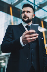 Fototapeta premium Below view of Caucasian government worker with cell smartphone device in hand looking away and pondering on financial news from content website, pensive intelligent expert with mobile phone