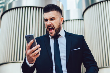 Crazy male entrepreneur feeling anger from trouble with solving enterprise problems shouting on cell smartphone gadget in financial district in megalopolis, unhappy man screaming to mobile gadget