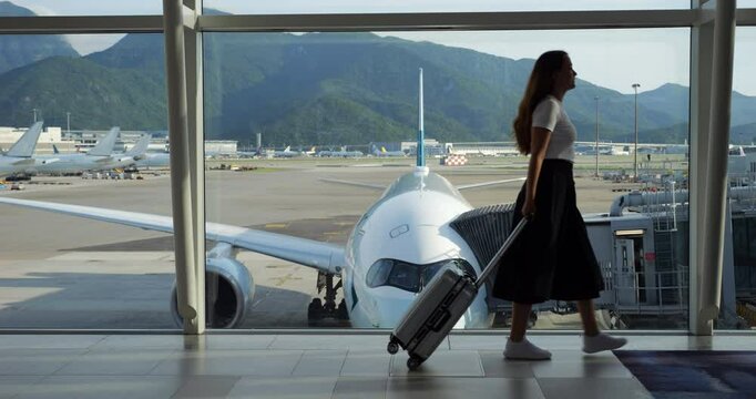 Passenger woman walks to boarding gate through empty airport lounge, pulling suitcase. Full-length slow motion shot shows her passing along window, parked airliner and airport grounds on background