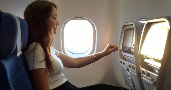 Woman sits in economy class by window, interacting with touchscreen of in-flight entertainment system on seatback in front of her. She selects movie to watch during long journey