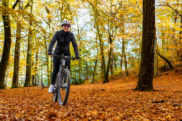 Middle aged woman wearing black sporty jacket, black cycling pants and white bike helmet riding bicycle in forest in autumn scenery. Front view.
