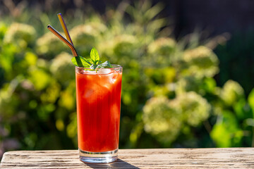 Bloody mary cocktail with ice cubes and green mint leaves on a nature background. Red tomato juice in transparent glass on a wooden table on a sunny day on the table, closeup