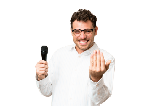Brazilian man picking up a microphone over isolated chroma key background inviting to come with hand. Happy that you came