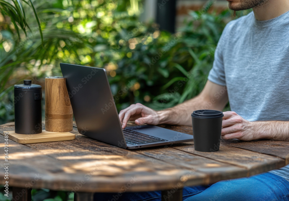 Poster a man sits at an outdoor table with a laptop and a mug of coffee, holding a black glass in his hand.