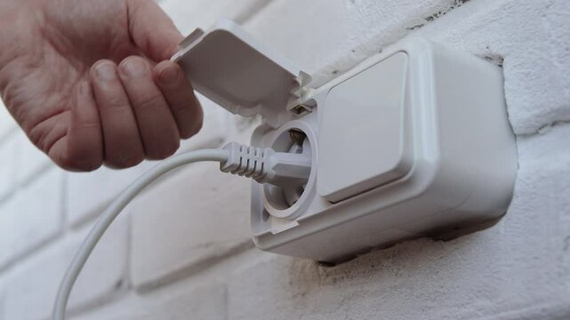 Hand inserting electrical plug into and plug out socket and on white wall close up, depicting energy use and power connectivity. Daily life, technology and household activities.