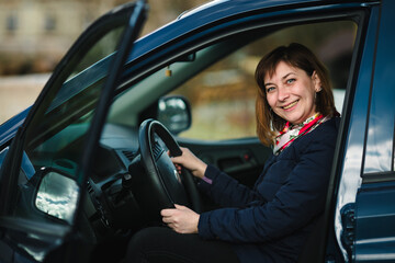 Naklejka premium A portrait of a woman sitting behind the wheel of her car, looking calmly at the camera with a soft smile. The car's interior is visible, and sunlight streams 