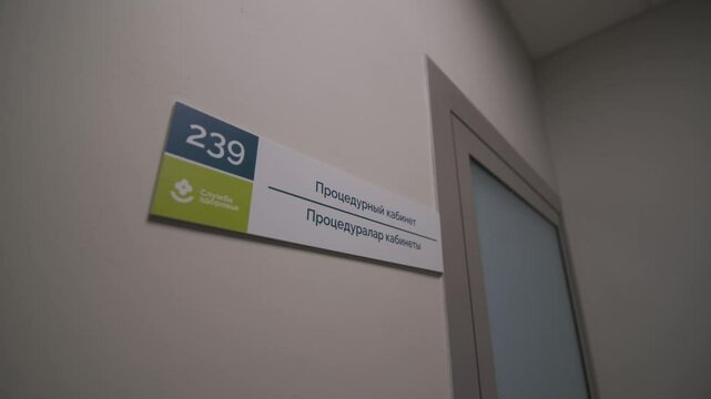 Treatment room nameplate with number on wall in hallway. Information for patients near office door. Table with text and digits in hospital
