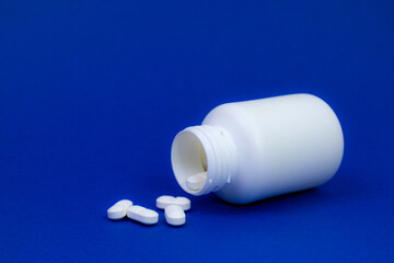 An open white pill bottle tipped over on its side against a blue background. A few white tablets have spilled out in front of the bottle. Healthcare, medication, pharmacy and wellness concept