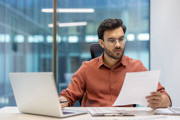 Businessman in office analyzing documents and working on laptop. Focused professional handling tasks with efficiency and concentration. Corporate environment facilitating dedicated workday activities