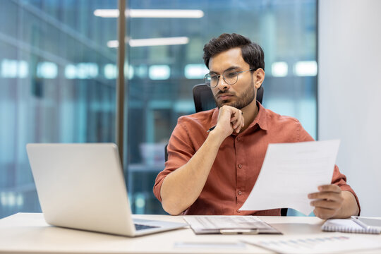Serious businessman thinking while reviewing document at office desk. Professional wearing glasses focused on paperwork with laptop in front. Calm environment reflects dedication, concentration