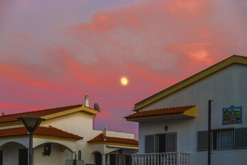 Moonrise over rooftops with a vibrant pink and orange sunset sky.