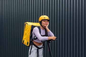 Bored Latin food courier with yellow backpack and helmet leaning on electric scooter