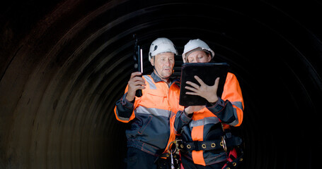 A couple of senior civil engineers are collaborating in the tunnel of a sewage drain, using a...