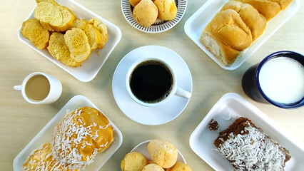 Cup of coffee in the center of breakfast table countaining cheese bread, butter toast, sweet bread, sweet cake with grated coconut on top, cup of milk and cup of latte or coffee with milk.
