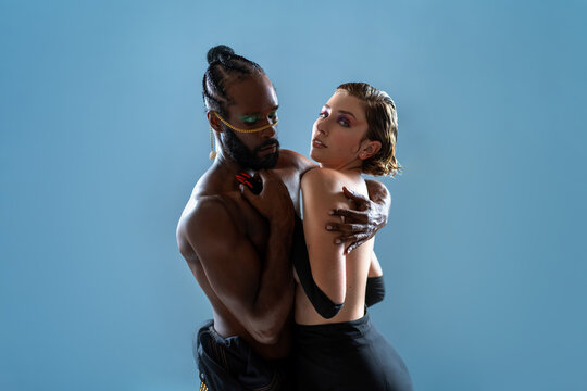 Studio shot of two LGBT people hugging in dance pose. Black gay man wearing bright blue make up holding girlfriend dancing isolated on blue background. - Powered by Adobe