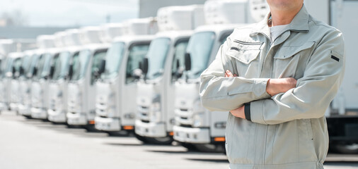 物流センターの駐車場に並ぶトラックの前にいる作業服姿の腕組みする男性（配送業・運送業・宅配業）  © buritora