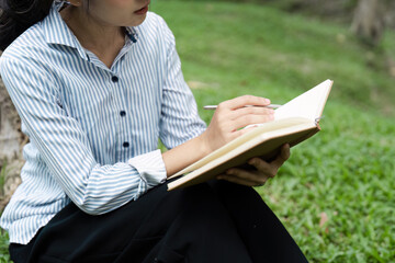 Young Woman Writing in Notebook While Sitting on Grass in Park, Relaxing Outdoors, Wearing Striped Shirt and Black Pants