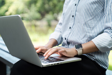 Fototapeta premium Close-up of Person Typing on Laptop Outdoors in a Park Setting, Wearing a Striped Shirt and Smartwatch