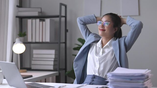 Businesswoman relaxing at her desk stretches her arms behind her head, enjoying a break from work. A bright, organized office enhances her wellbeing and promotes stress relief