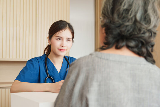 Southeast Asian nurse in blue uniform listening attentively to elderly patient at clinic reception desk, highlighting trust, communication, and compassionate healthcare service.