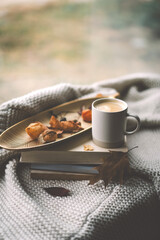 A warm cup of coffee sits atop a golden tray adorned with dried leaves, resting on a stack of books, with a cozy blanket draping nearby.