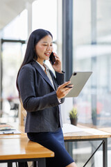 Fototapeta premium Modern business asian woman in formalwear using digital tablet while standing near wooden desk in an office interior in the office. business people concept.