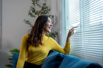 Young woman sitting on sofa is opening blinds and looking through the window