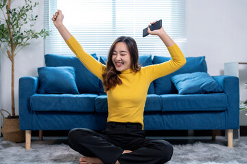 Young asian woman celebrating success while holding smartphone at home