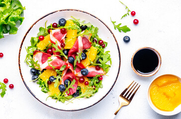 Delicious salad with smoked duck fillet, juicy orange, fresh blueberries, cranberries and arugula on white background, top view