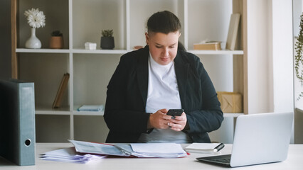 Lazy employee. Office procrastination. Bored distracted overweight woman lacking motivation typing in chat playing phone ignoring documents paperwork.