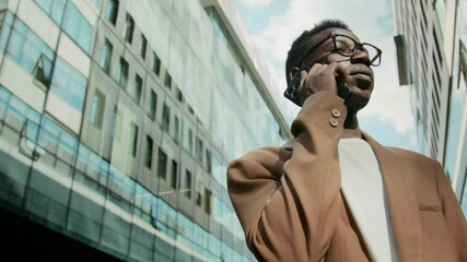 Low angle shot of Black young businessman dressed formally speaking on phone between large office buildings