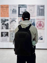 Young man with a cap and jacket watching different paintings while carrying a backpack and a green bottle.