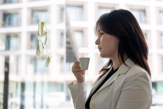 A businesswoman is enjoying her coffee while reading ideas on sticky notes attached to a glass wall.