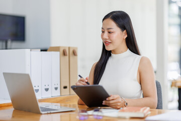 Positive young asian female freelancer in formal suit sitting at table with laptop and browsing tablet while working on project in creative workspace in daylight