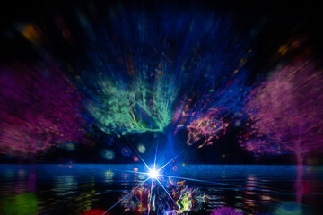 A mesmerizing light projection of trees in vivid colors illuminates a dark scene, with reflections on the water and a bright starburst at the base. The display radiates vibrant energy and surreal