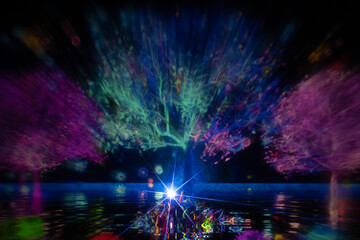 A mesmerizing light projection of trees in vivid colors illuminates a dark scene, with reflections on the water and a bright starburst at the base. The display radiates vibrant energy and surreal