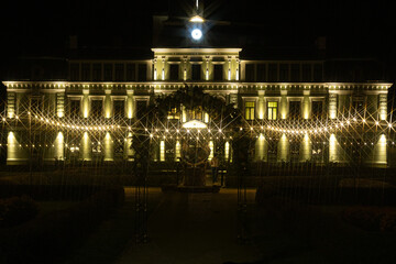 A grand view of Koceni Manor at night, beautifully lit with golden lights that create a starburst...