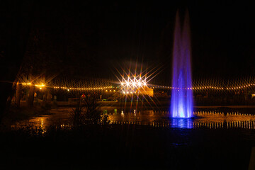 A vibrant blue fountain glows in the night, surrounded by reflections on the water and festive string lights. Warm golden lights and starbursts enhance the serene nighttime scene.