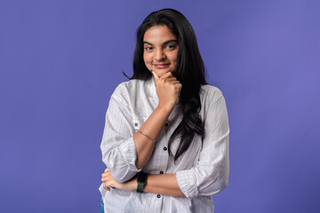 A young woman of Indian descent, wearing a white striped shirt and a black smartwatch, stands with a hand on her chin, exuding confidence and contemplation against a solid purple background