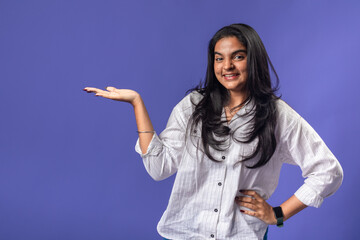 A young woman of Indian descent, wearing a white striped shirt and black smartwatch, stands against a solid purple background, smiling and gesturing with her palm up