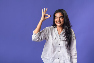 A young woman of Indian descent, wearing a white striped shirt and black smartwatch, stands against...