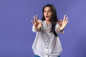 A young woman of Indian descent, wearing a white striped shirt and black smartwatch, stands against...