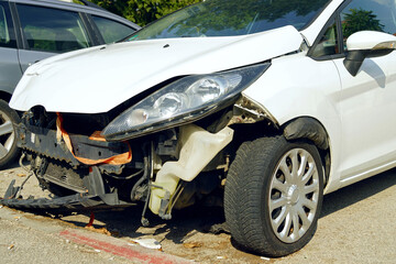 A close-up photo of the front of a white passenger car with a torn off bumper. The aftermath of a car accident on the road: damage to vehicles after a head-on collision.