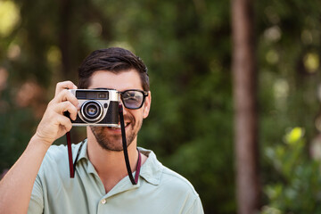 A man smiles while taking a photo with a vintage camera in a sunny park. He enjoys the outdoors, surrounded by greenery, in a relaxed and playful moment. 